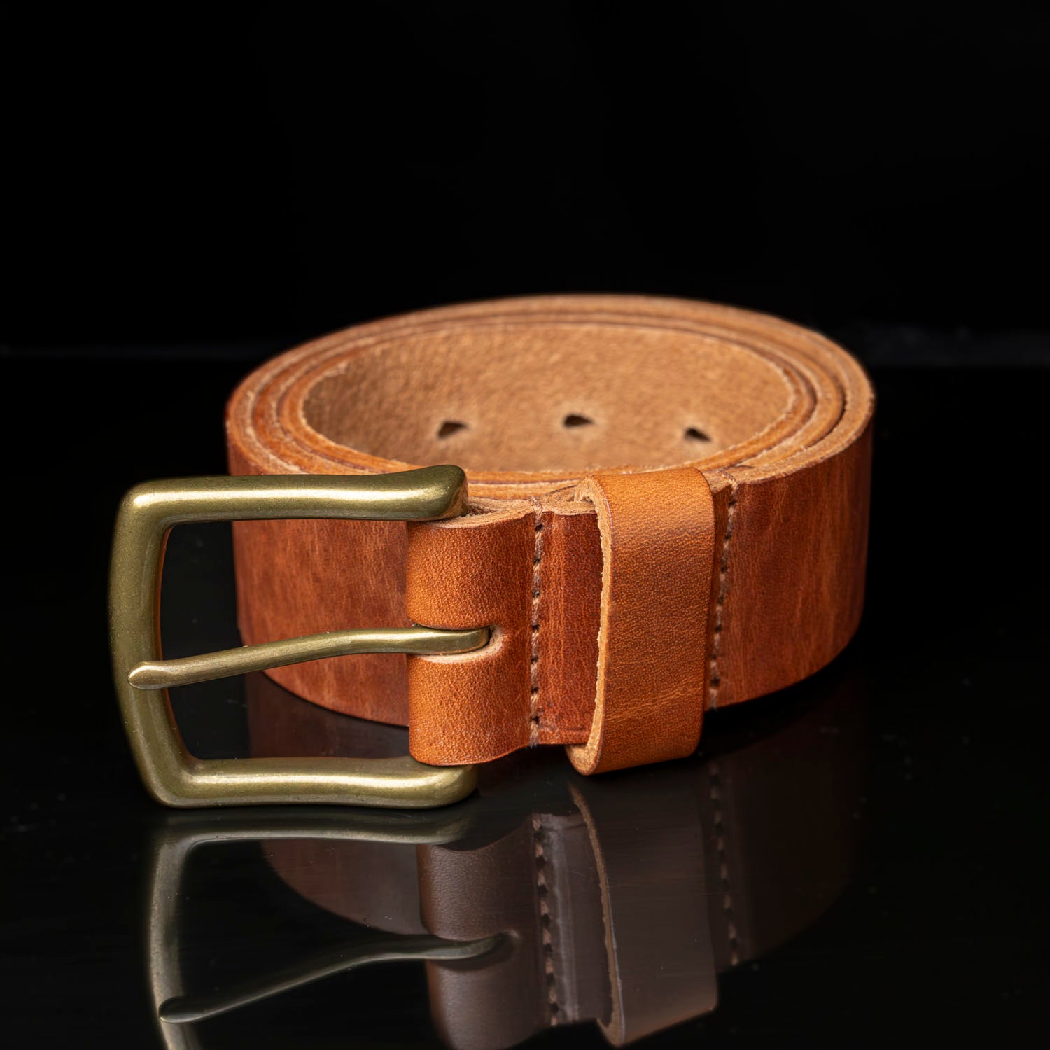 A brown leather belt with a solid brass buckle and seven tear-drop adjustment holes.