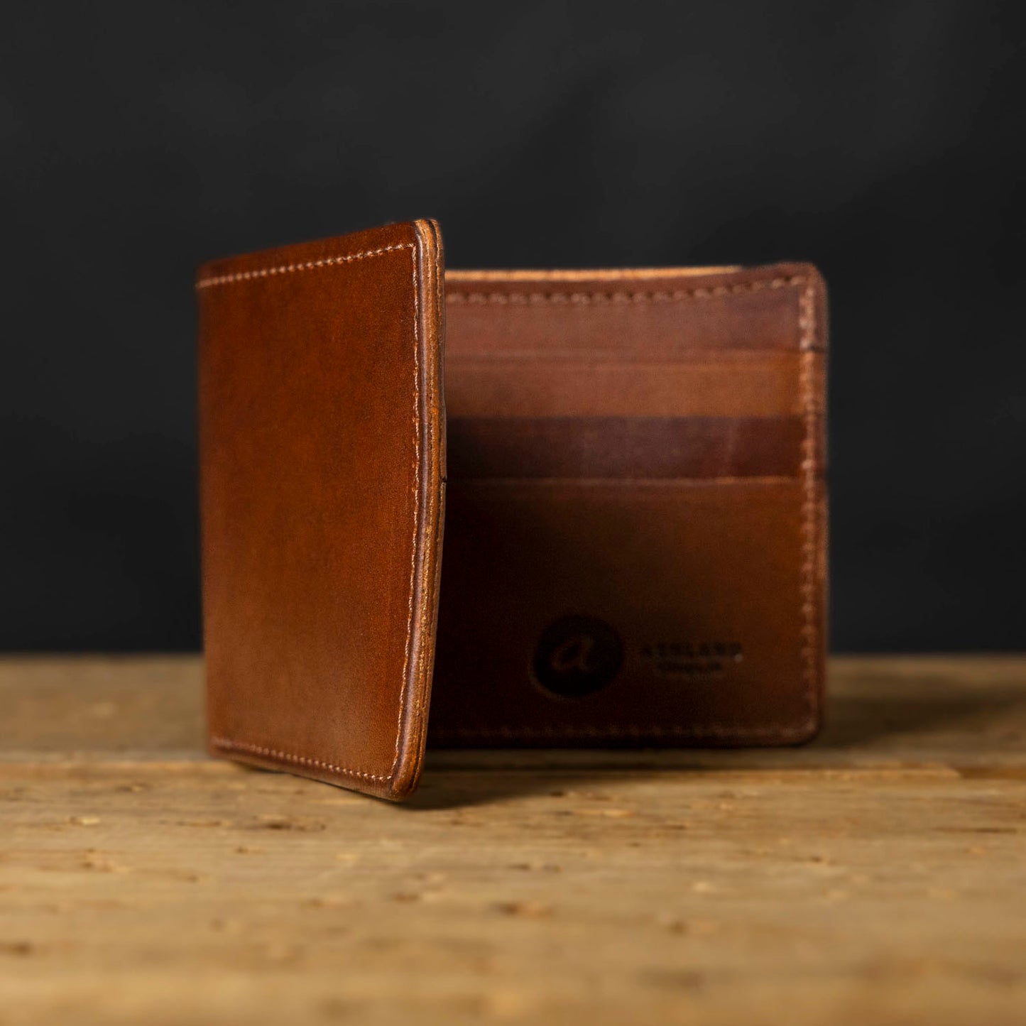 A brown bifold leather wallet with visible stitching and a logo embossed on the bottom right. The wallet is displayed in an open and closed position on a wooden surface.
